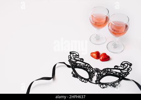 Valentines Day Festive table setting with black lace mask, two glasses of rose wine and two red heart shape candies on white table. Valentine Day Stock Photo