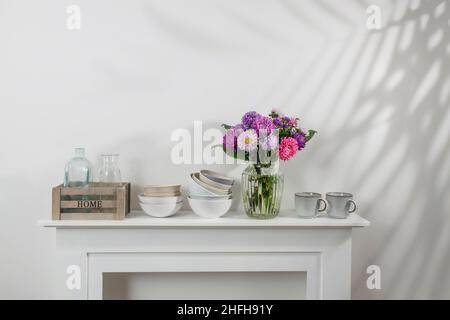 kitchen utensils, cups, bowls, bottles, storage items, a bouquet of asters are on a white dresser. Copy space Stock Photo
