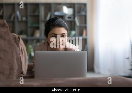 Pleasant happy young Indian woman using computer, resting on sofa. Stock Photo