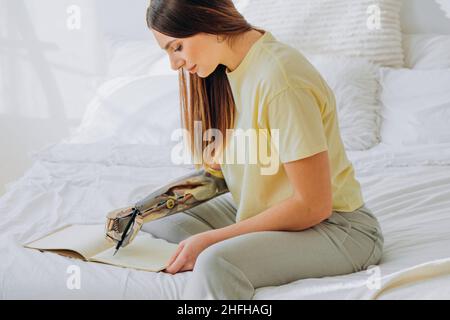 Young woman in yellow t-shirt writes in paper notebook with bionic arm sitting on bed in contemporary room at home closeup Stock Photo