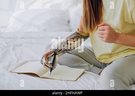 Young woman in yellow t-shirt writes in paper notebook with bionic arm sitting on bed in contemporary room at home closeup Stock Photo