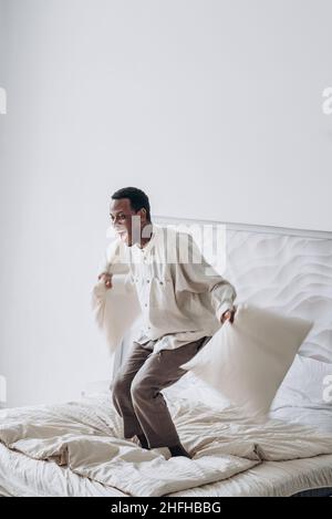 Joyful young African-American man in casual clothes has fun jumping and throwing pillows on large bed at home in sunny morning Stock Photo