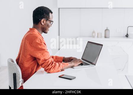 Concentrated young African-American guy in orange clothes and glasses works on modern laptop at comfortable workplace closeup Stock Photo