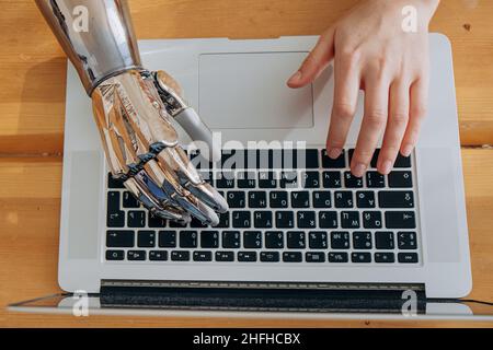 Young woman uses modern metal bionic arm to type on laptop keyboard sitting at wooden table in light room extreme close view Stock Photo