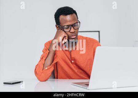 Tired young African-American man with glasses sleeps leaning on hand near laptop at white table in light room close view Stock Photo