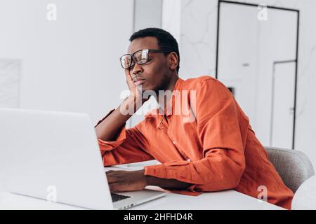 Tired young African-American man with glasses sleeps leaning on hand near laptop at white table in light room close view Stock Photo