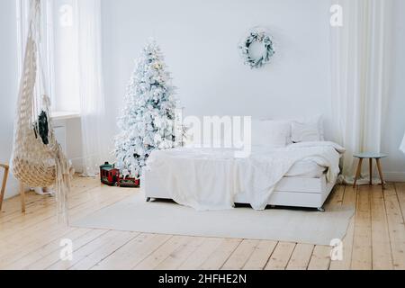 A bright, spacious room with a large bed with white sheets and an elegant light Christmas tree and a wreath on the wall. Cute and festive interior. Stock Photo
