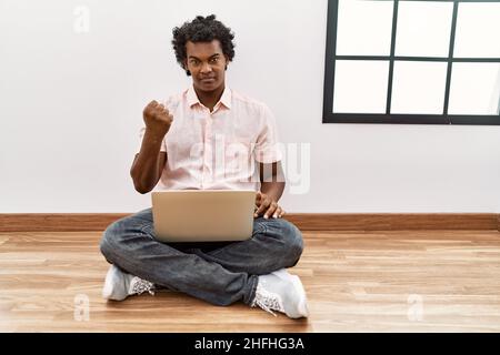 African man with curly hair using laptop sitting on the floor angry and mad raising fist frustrated and furious while shouting with anger. rage and ag Stock Photo