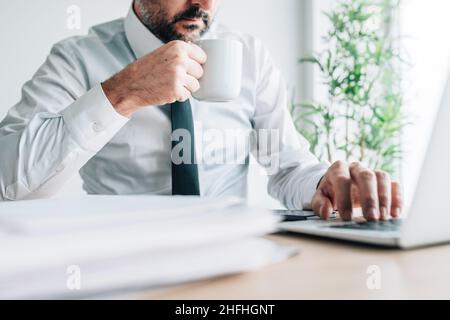 Drinking coffee at work increases productivity, businessman with cup of hot beverage working on laptop computer, selective focus Stock Photo
