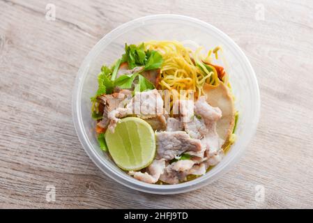 Dry noodles pork with red roasted pork and vegetable in plastic bowl on wooden table, top view Thai and China Asian food yellow noodles lemon lime Stock Photo
