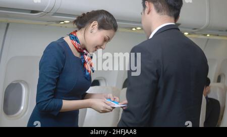 Flight attendant woman or Air hostess in uniform service take care of male passengers checking seats boarding pass and passport on the airplane before Stock Photo