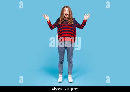 Full length portrait of surprised woman opening her mouth and raised arms in amazement, hearing awesome news, pleasantly surprised by sudden success. Indoor studio shot isolated on blue background. Stock Photo