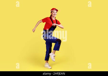 Full length portrait of optimistic positive worker woman marching, posing on one leg, going to do her work, wearing overalls and red cap. Indoor studio shot isolated on yellow background. Stock Photo