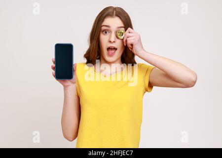 Surprised beautiful teenager with brown hair in yellow T-shirt holding mobile phone and covering eye with bitcoin, shocked about cryptocurrency apps. Indoor studio shot isolated on gray background. Stock Photo