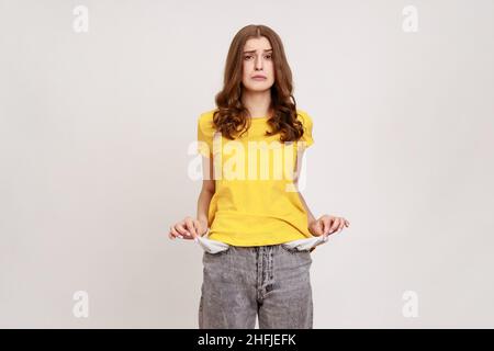 Portrait of frustrated worried brown haired female of young age in casual yellow T-shirt turning out empty pockets showing no money gesture. Indoor studio shot isolated on gray background. Stock Photo