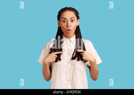 Astonished young woman with black dreadlocks pointing at herself, asks who me, has surprised expression, shocked being picked, wearing white shirt. Indoor studio shot isolated on blue background. Stock Photo