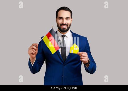 Satisfied man holding German flag and paper house, dreaming to buy his own house or flat in Germany, looking at camera, wearing official style suit. Indoor studio shot isolated on gray background. Stock Photo