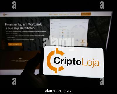 Person holding smartphone with logo of Portuguese company Smart Token Lda. (CriptoLoja) on screen in front of website. Focus on phone display. Stock Photo