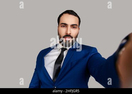 Portrait of handsome bearded man taking selfie, looking at camera with relaxed optimistic calm emotions, POV, wearing official style suit. Indoor studio shot isolated on gray background. Stock Photo