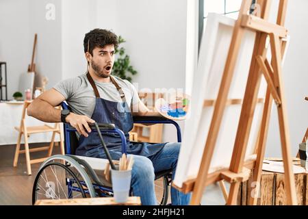 Young hispanic man sitting on wheelchair painting at art studio in shock face, looking skeptical and sarcastic, surprised with open mouth Stock Photo