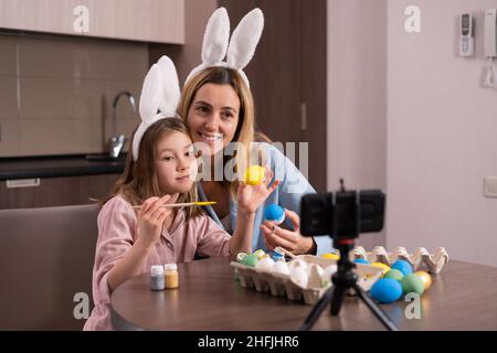 caucasian mother and daughter in a bandage on the ears of a rabbit are sitting in the kitchen at home. decorate Easter eggs take a selfie using a smar Stock Photo