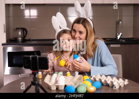 caucasian mother and daughter in a bandage on the ears of a rabbit are sitting in the kitchen at home. decorate Easter eggs take a selfie using a smar Stock Photo
