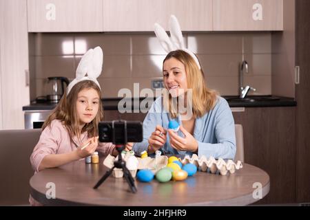 mother and daughter in a bandage on the ears of a rabbit are sitting in the kitchen at home. They decorate Easter eggs with paints and take selfies on Stock Photo