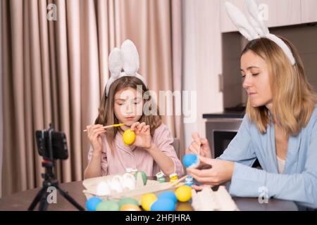 caucasian mother and daughter in a bandage on the ears of a rabbit are sitting in the kitchen at home. decorate Easter eggs with paints and video call Stock Photo