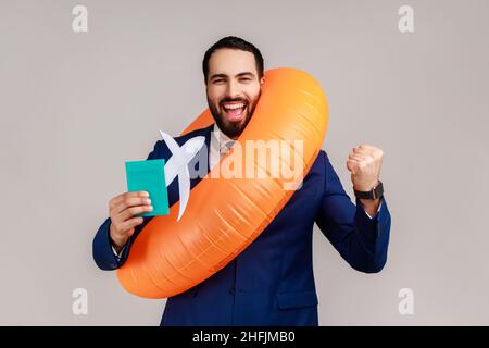 Happy bearded businessman holding orange rubber ring, paper plane and passport, clenching fist, long awaited vacation, wearing official style suit. Indoor studio shot isolated on gray background. Stock Photo