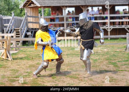 Two knights are fighting in armor with swords, reconstruction of the tournament. A scene with spectators from a log house. Celebration of the pagan da Stock Photo