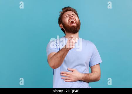 Hilarious laughter. Portrait of joyful happy bearded man laughing loudly and pointing to camera, mocking taunting you, holding belly. Indoor studio shot isolated on blue background. Stock Photo