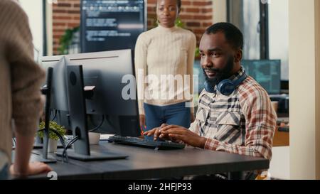 Relaxed software developer wearing wireless headphones smiling working looking at computer screen while typing. Coder writing code in it agency office with colleague asking for informations. Stock Photo