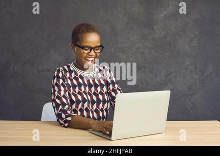 Positive african american having fun laughing while sitting at laptop on gray background. Stock Photo