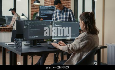 Focused software developer writing code looking at multiple computer screens displaying machine learning algorithm. Programer coding user interface while colleagues doing teamwork in background. Stock Photo
