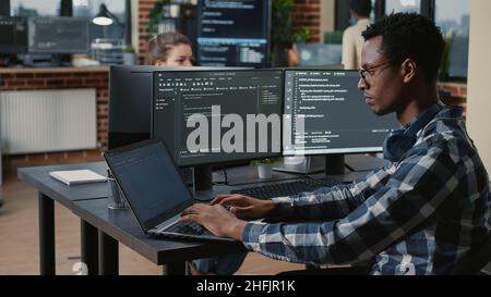 Portrait of tired programer rubbing eyes while typing on laptop sitting at desk with multiple screens parsing code in software agency. Coder losing focus while working on user interface. Stock Photo