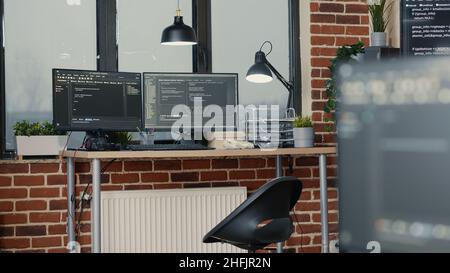 Desk with multiple computer monitors displaying parsing code and programming language compiling in empty software developing agency office. Servers cloud computing big data algorithms. Stock Photo