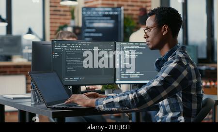 Portrait of african american developer using laptop to write code sitting at desk with multiple screens parsing algorithm in software agency. Coder working on user interface using portable computer. Stock Photo