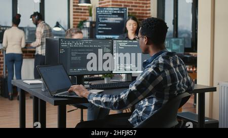 Portrait of system engineer fixing glasses while typing on laptop smiling satisfied about compiling results sitting at desk with multiple screens. App developer enjoying working on user interface. Stock Photo