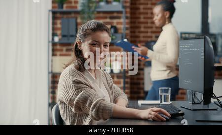 Programer typing source code on computer keyboard turning head and smiling at camera. Casual software engineer sitting at desk in it startup company writing ai algorithm feeling confident. Stock Photo