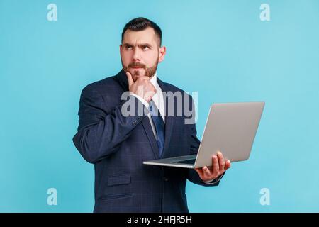 Portrait of pensive smart businessman wearing dark official style suit holding laptop and thinking about new project, holding his chin. Indoor studio shot isolated on blue background. Stock Photo