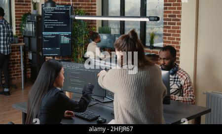 Software programer pointing pencil at source code on computer screen explaining algorithm to coworker standing next to desk. Programmers discussing online cloud computing with team. Stock Photo