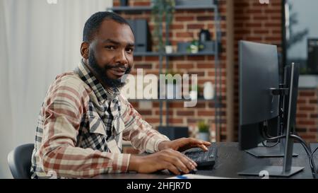 Portrait of programer typing programming code on computer keyboard turning head and smiling while colleague walks by in background. Software engineer sitting at desk writing code feeling confident. Stock Photo