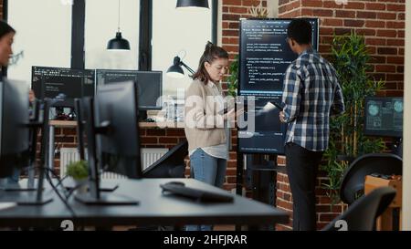 Software developer holding digital tablet analyzing code on wall screen tv explaining errors to coworker programer in busy office. Team of coders collaborating on ai cyber security group project. Stock Photo