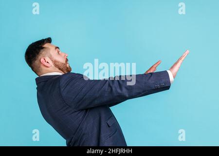 No, it's scary. Profile portrait of frightened shocked man in suit raising hands in fear, looking horrified and panicking, hiding from phobia. Indoor studio shot isolated on blue background. Stock Photo