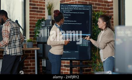 Programmer holding digital tablet analyzing code on wall screen tv while colleague is looking for errors pointing using remote control. Team of app developers collaborating on big data group project. Stock Photo