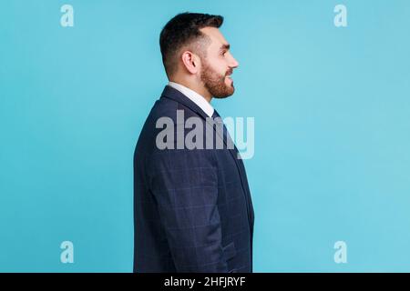 Profile portrait of young adult handsome bearded businessman wearing official style suit looking ahead with toothy smile, optimism, success. Indoor studio shot isolated on blue background. Stock Photo