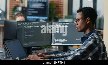 Sofware developer thinking while touching beard while typing on laptop sitting at desk with multiple screens parsing code. Focused database admin working with team coding in the background. Stock Photo