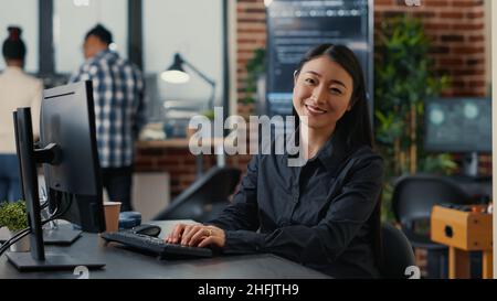 Smiling asian database software developer writing programming code on computer keyboard sitting at desk in software development agency. Friendly cloud programer innovating cyber security. Stock Photo