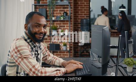 Database programer writing code and smiling while coworker walks by holding clipboard in it agency. Relaxed app developer typing on keyboard with colleagueas computing big data. Stock Photo