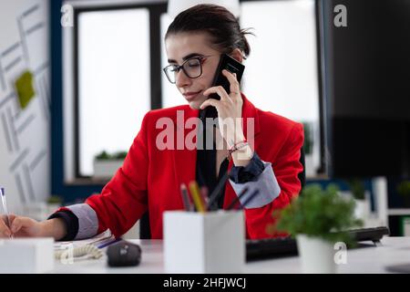 Businesswoman taking notes in smartphone phonecall sitting at desk. Entrepreneur with glasses working in startup office. Small business owner in red jacket writing on paper in startup enviroment. Stock Photo